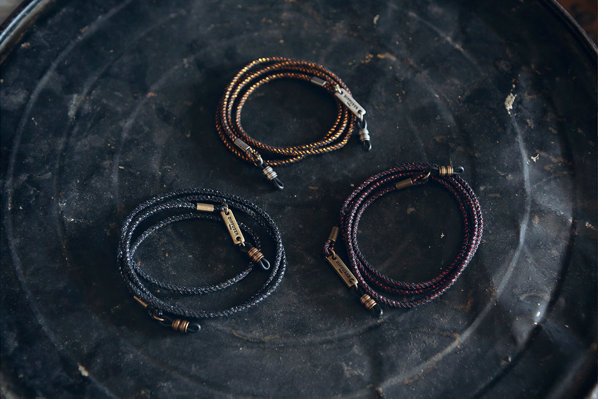 Three braided leather bracelets with brass-toned hardware are arranged on a dark textured surface. One bracelet is brown, one is black, and one is dark purple.