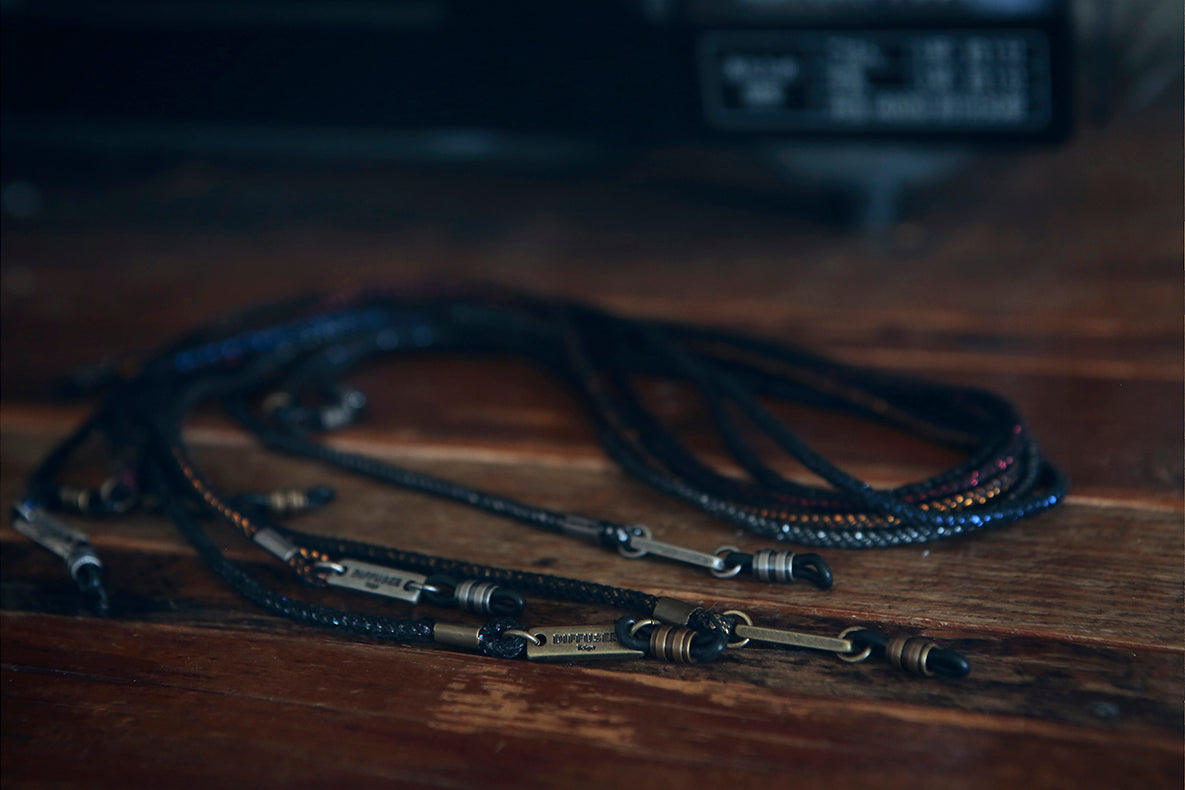 A set of dark braided necklaces with antique bronze-toned metal charms and rings, arranged on a rustic wooden tabletop with a blurred dark background.