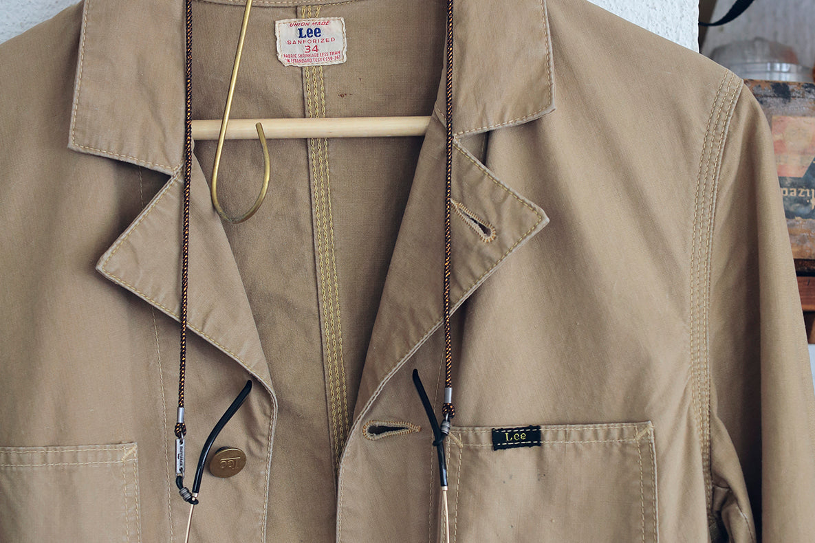 A beige Lee utility jacket hanging on a wooden hanger, featuring a pointed collar, front button closure, and a small black Lee brand patch on the chest pocket.