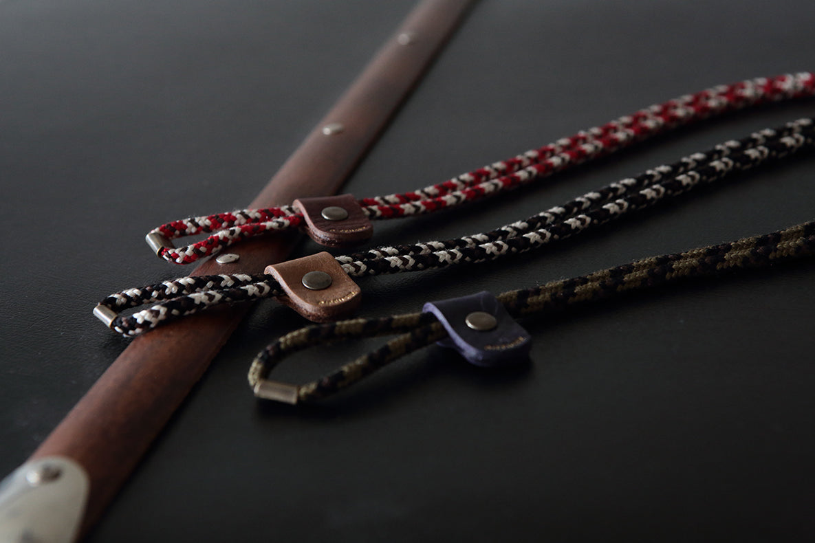 Three braided leather and cord lanyards with metal snap closures are arranged diagonally on a dark surface next to a wooden rod. The cords are red and white, black and white, and black and brown.