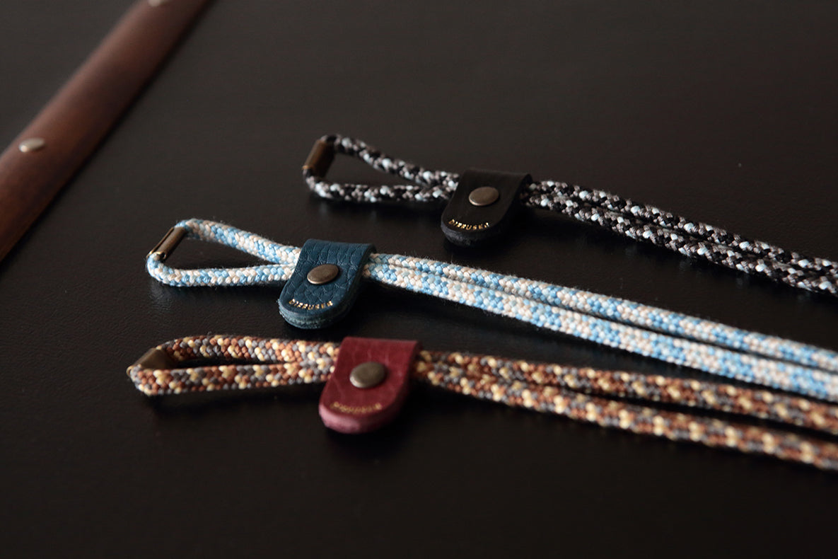 Three woven leather strap accessories with snap closures are arranged diagonally on a dark surface. The top strap is black and white, the middle is blue and white, and the bottom is brown and beige.