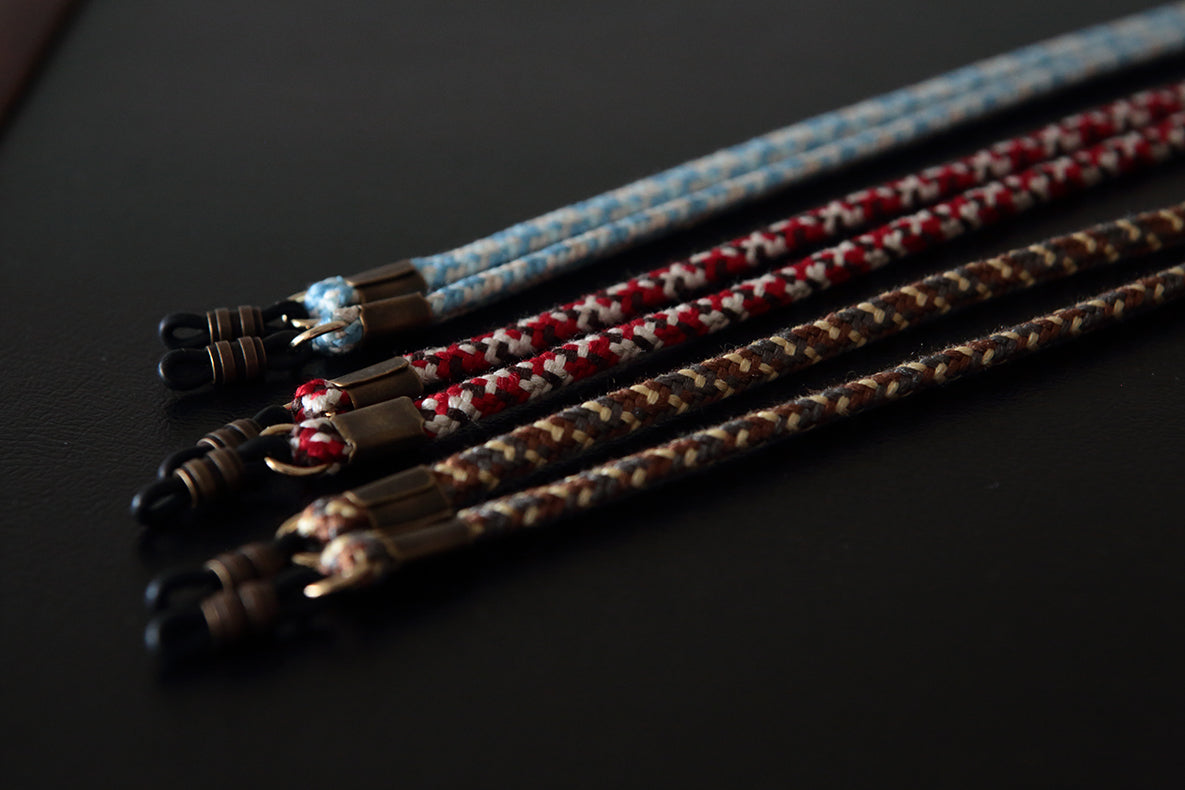 Three braided cord accessories with metal connectors, arranged diagonally on a dark surface. The cords are blue and white, red and white, and brown and beige.