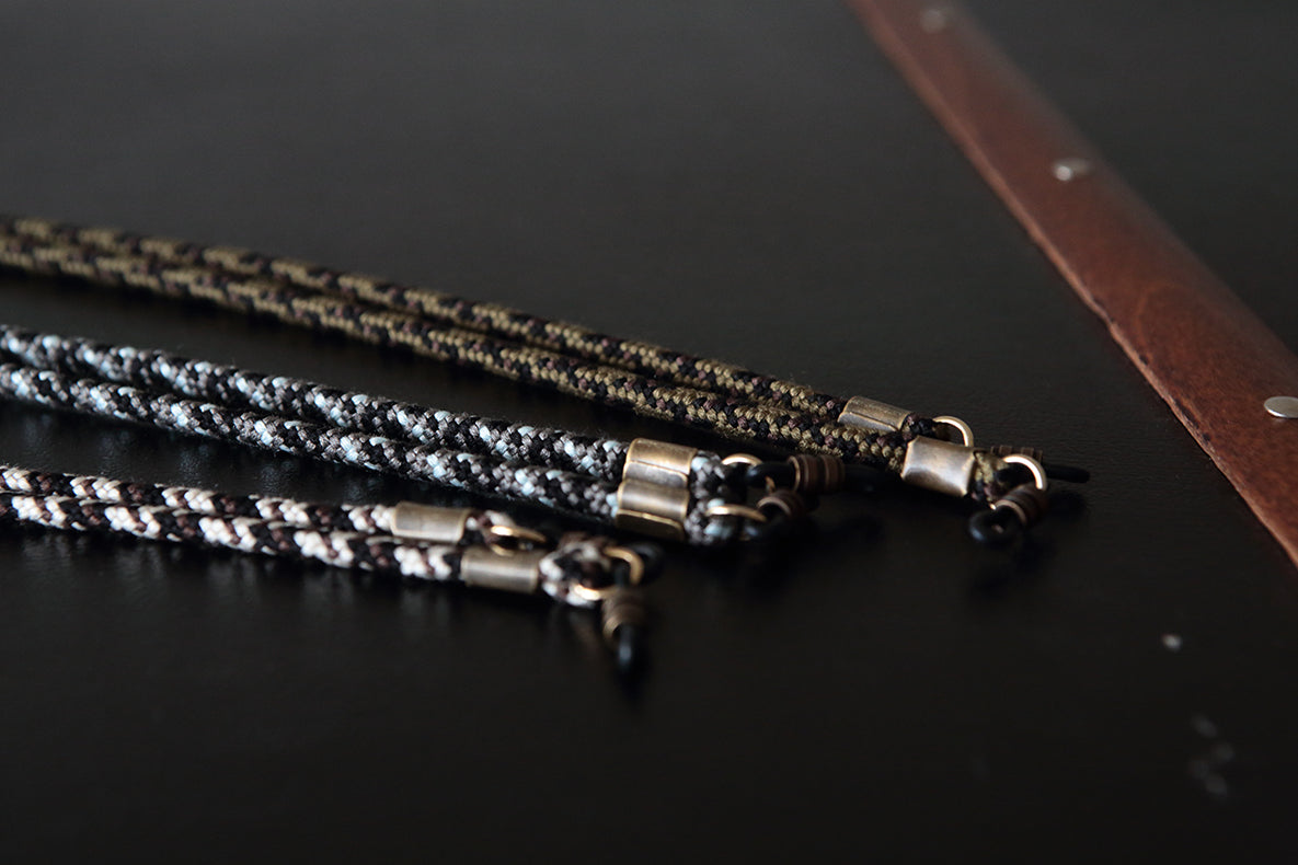 Three braided leather necklaces with silver-toned metal clasps arranged diagonally on a dark surface, with a brown leather strap visible in the upper right corner.