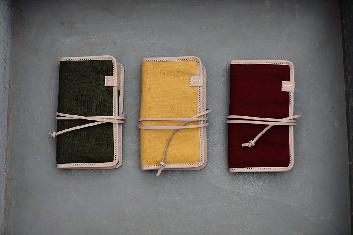 Three canvas notebook covers in dark green, mustard yellow, and burgundy, each wrapped with a beige cord and bordered with light beige stitching, arranged on a gray background.