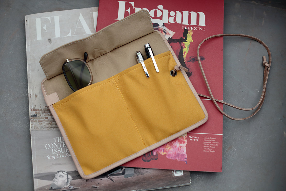 A mustard yellow canvas roll-top pouch with beige trim, shown open and styled on top of two magazines. The pouch has multiple pockets, including one holding sunglasses and two pens, and includes a brown leather strap.