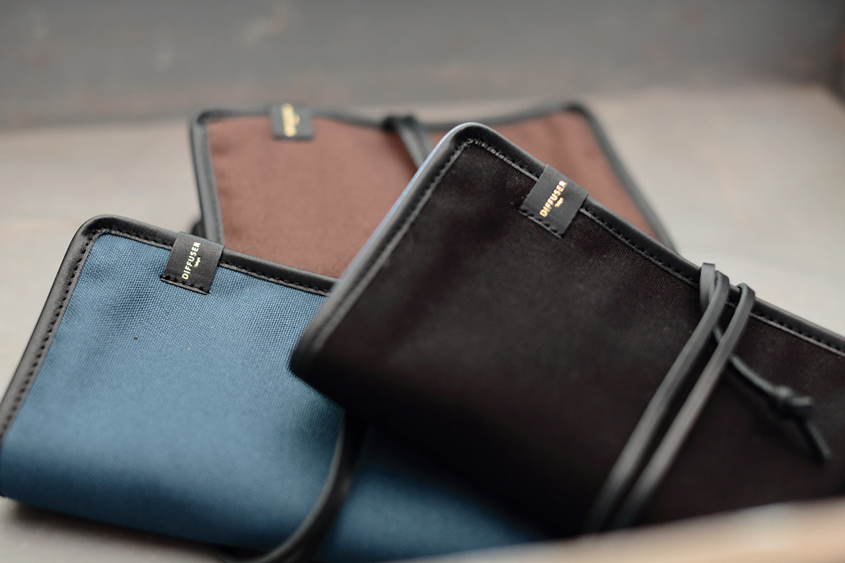 Three fabric document pouches with black trim and drawstring closures, shown overlapping on a neutral surface. The pouches are in blue, brown, and black, each with a small black brand tab reading 'SUGAR CANE'.