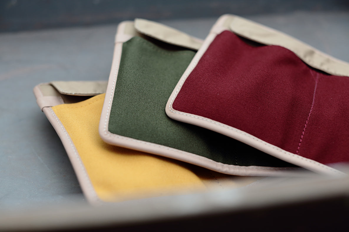 Three square fabric pouches or small mats in yellow, green, and burgundy with cream-colored stitched edges, arranged overlapping on a dark gray surface.