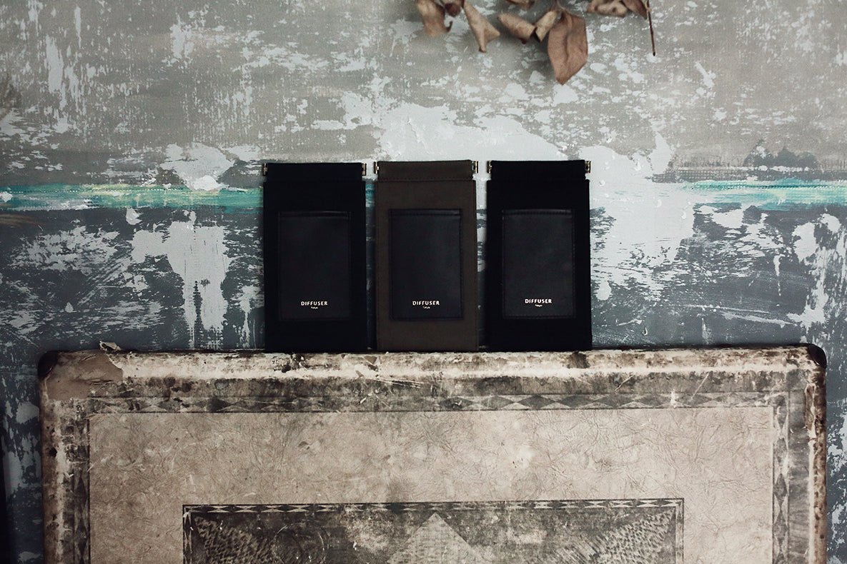 Three slim rectangular black and dark brown leather accessories with gold 'BUTTERFLY' branding, displayed side-by-side on a distressed concrete ledge against a textured gray wall.