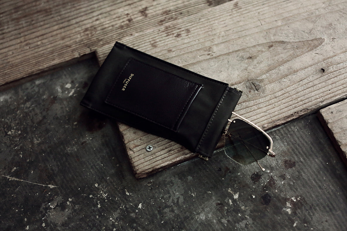 A black leather card holder wallet with a gold-embossed logo, shown with a pair of sunglasses on a rustic wooden and concrete surface.