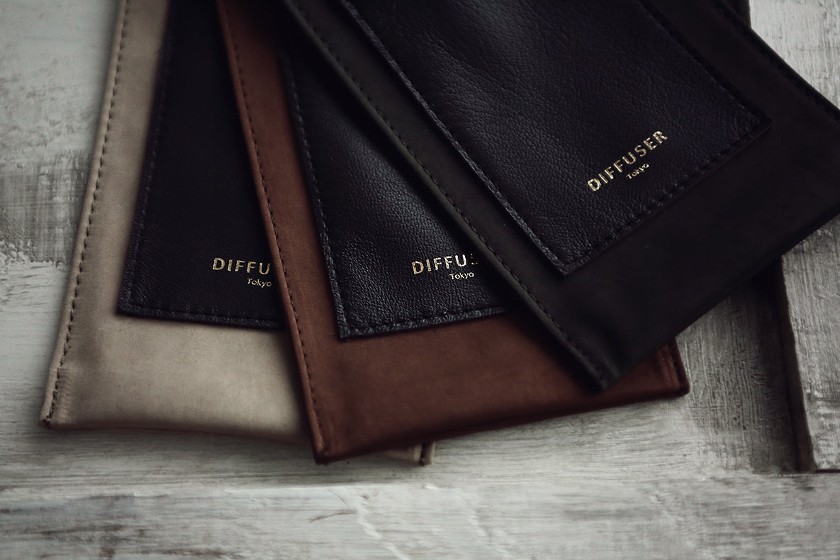 Four leather diffuser pouches in beige, black, brown, and dark gray, arranged diagonally on a rustic gray wooden surface. Each pouch features a stitched pocket with the brand name 'DIFFUSER London' in gold lettering.