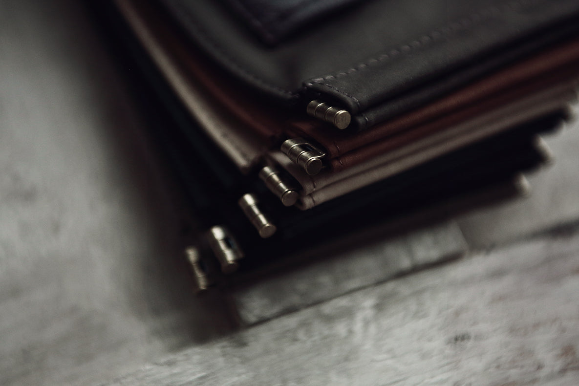 A close-up, angled view of a stack of leather wallets or card cases in dark brown and black, showing silver-toned metal snap buttons, resting on a textured gray surface.