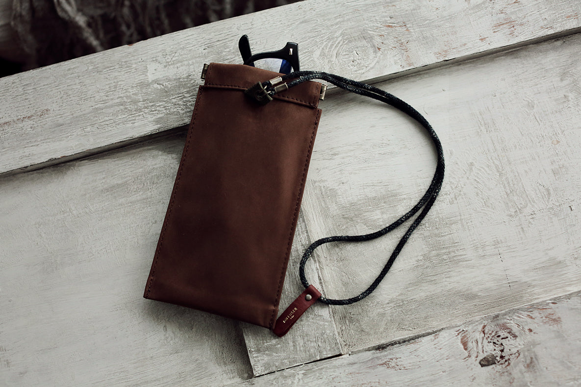 A brown leather pouch with a black strap and black metal hardware, photographed resting on a weathered white wooden surface.