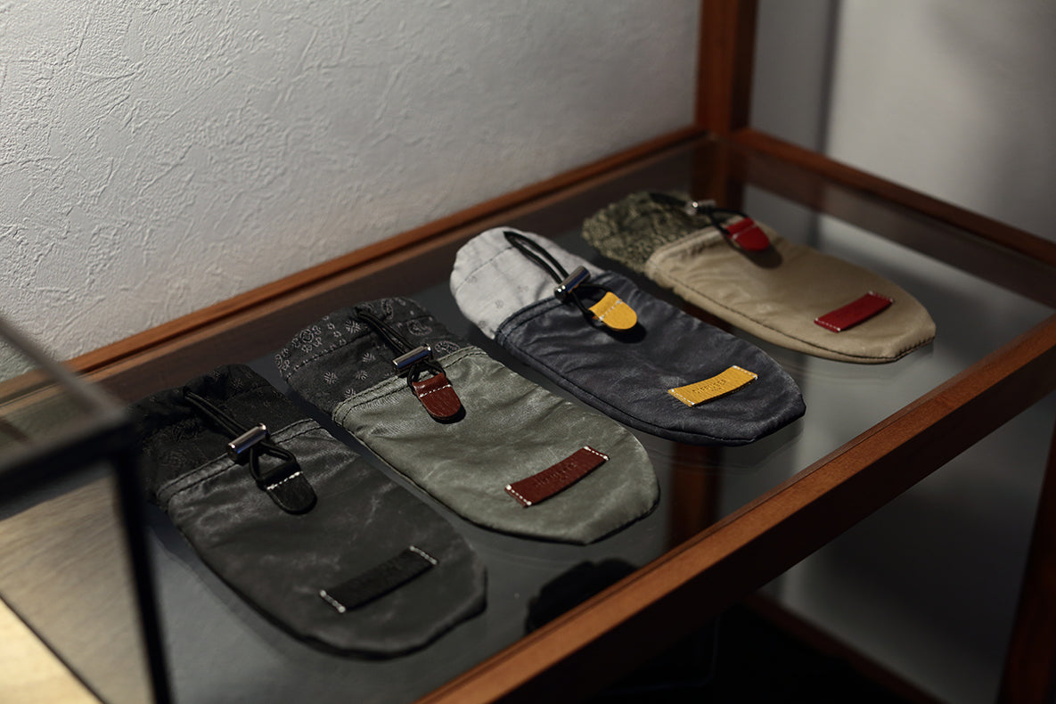 Four slip-on loafers in different colors displayed on a glass shelf inside a wooden case. The shoes include dark gray, olive green, charcoal, and beige, each with a leather strap and a small leather accent tab.