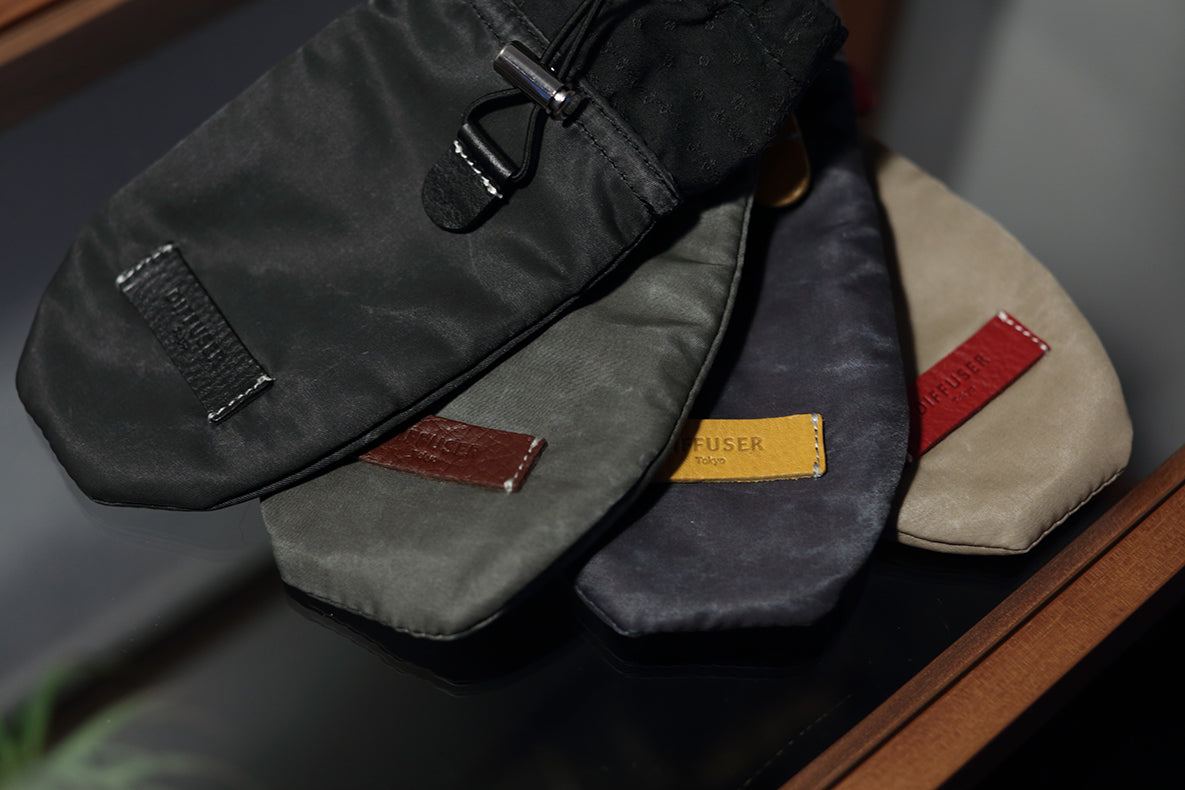 Four fabric pouches or small bags in different colors arranged on a dark surface: black, dark gray, charcoal, and beige, each with a small leather patch and zipper detail.