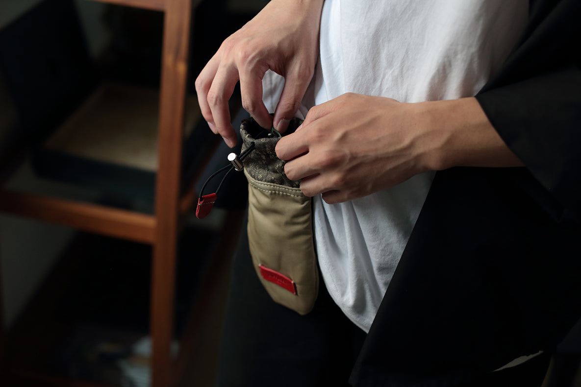 A close-up of a person's hands opening a beige canvas pouch with a black zipper and a small red tab, worn over a white t-shirt and black jacket.
