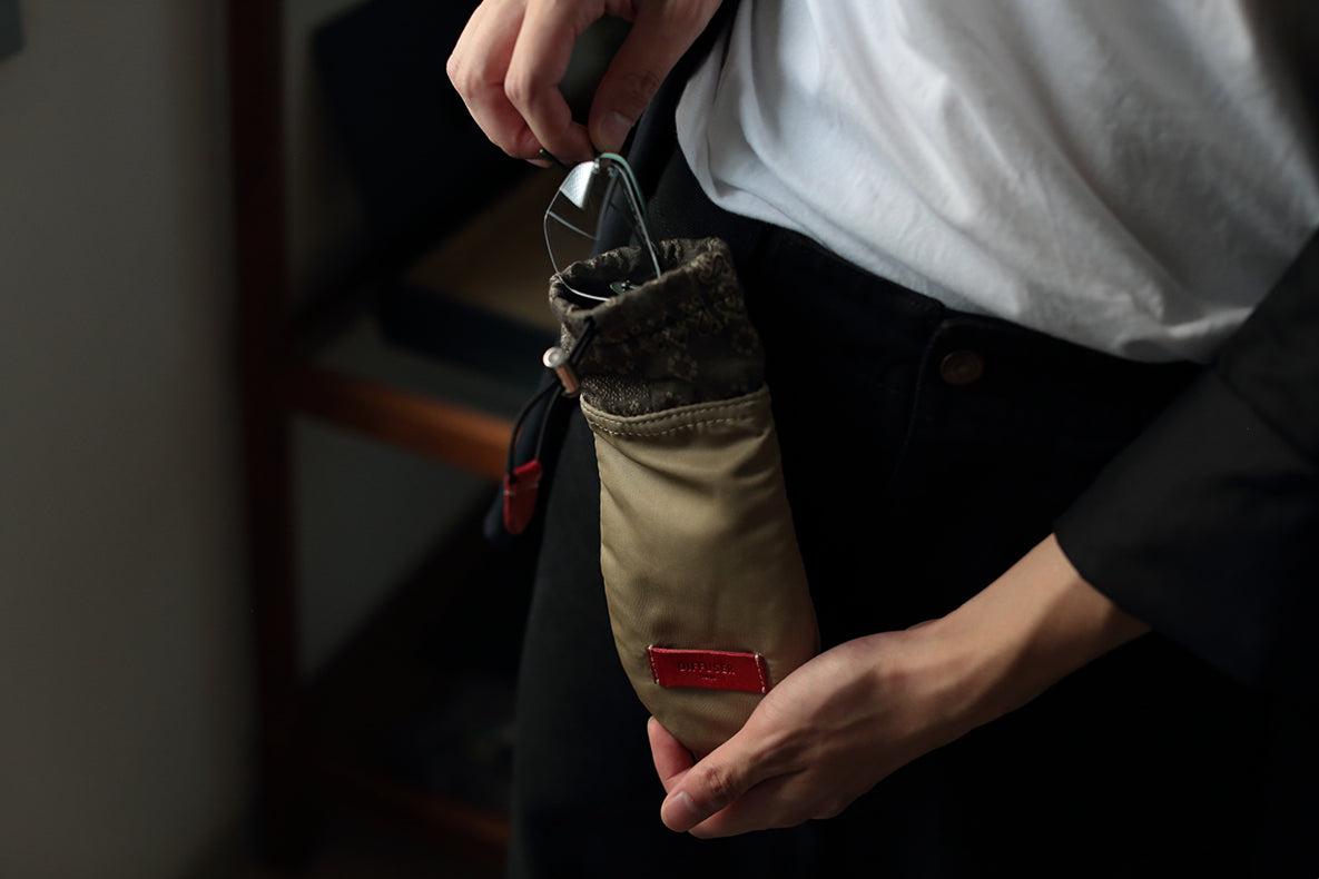 A close-up of a person holding a beige canvas pouch with a dark gray fuzzy trim and a small red label. The person is wearing a white shirt and black pants, and the pouch is shown in a real indoor environment.