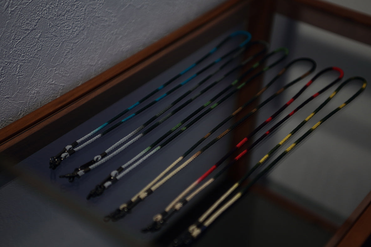 DIFFUSER STRIPE DYEING GLASS CODE lanyards by DIFFUSER, crafted from vibrant Japanese polyester cord and finished with metal clips, are neatly arranged on a glass display case framed in wood, set against a textured white wall and illuminated by soft lighting.