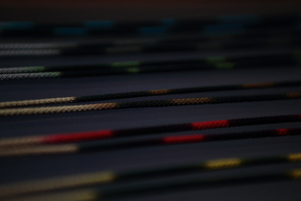 Close-up of several colorful, braided DIFFUSER STRIPE DYEING GLASS CODE cords by DIFFUSER—showcasing intricate dye patterns and craftsmanship—lying parallel on a dark surface, with selective focus creating a blurred, abstract effect.