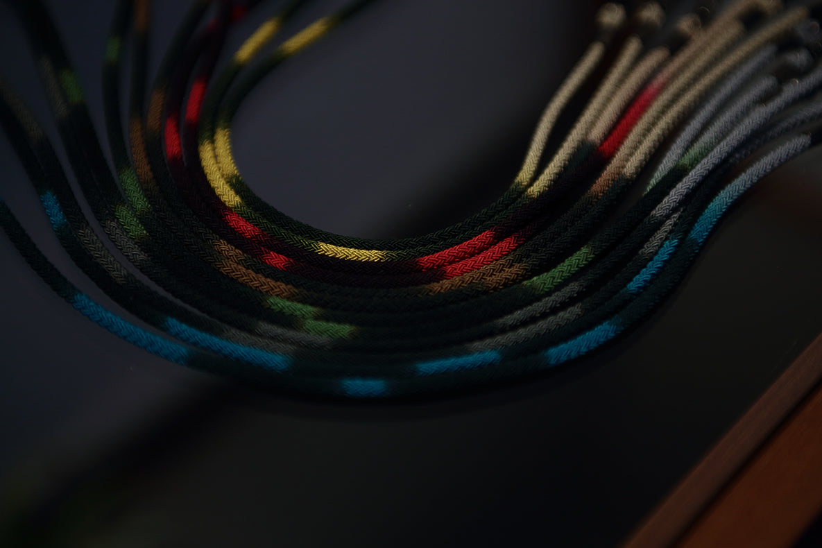 Several black DIFFUSER STRIPE DYEING GLASS CODES by DIFFUSER, crafted in Japan with traditional Kasuri dyeing techniques, are arranged in parallel on a dark surface. The polyester cords feature vibrant yellow, red, blue, and green patterns that stand out against the dimly lit background, highlighting the colorful threads.