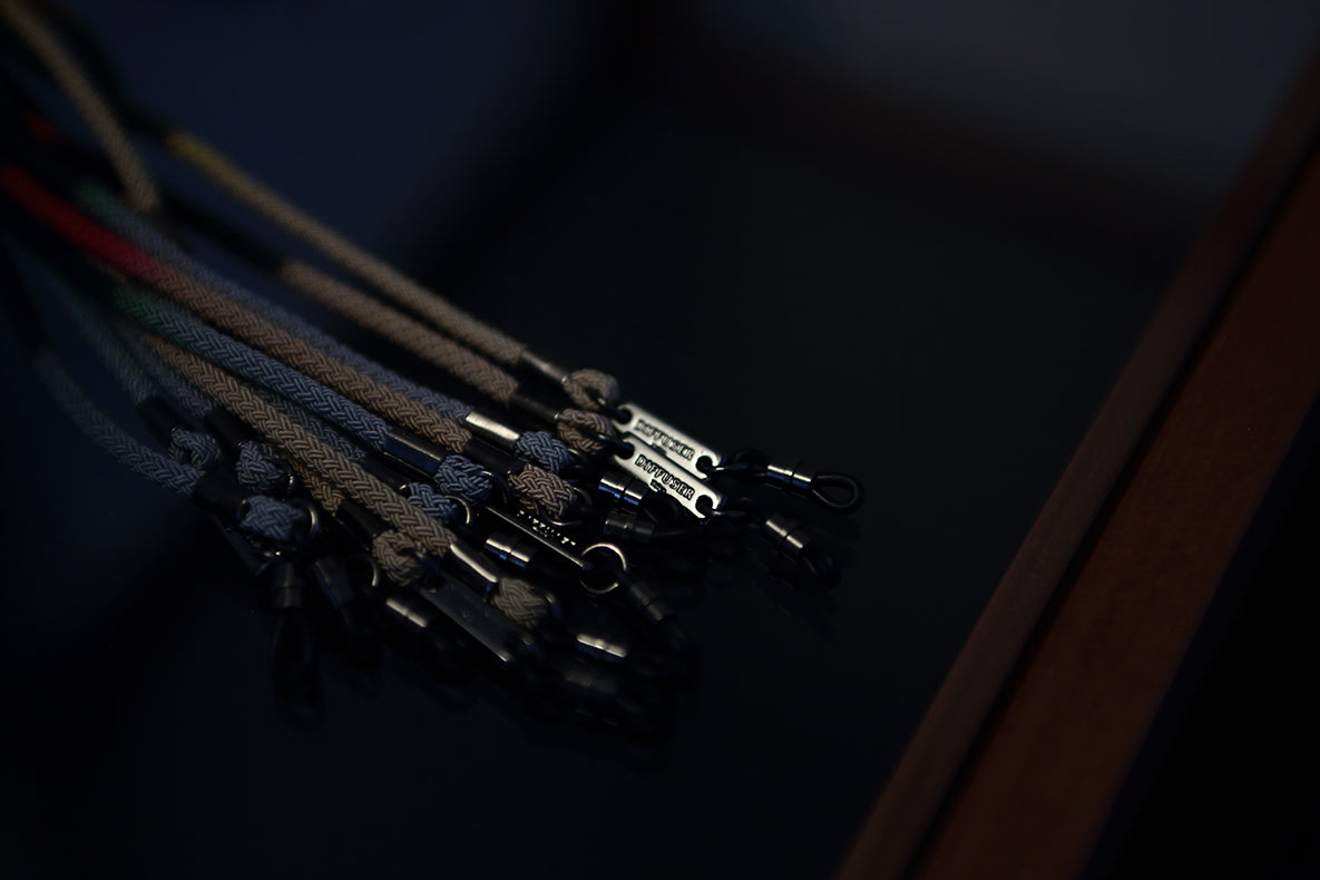 Several DIFFUSER STRIPE DYEING GLASS CODE lanyards by DIFFUSER, braided from Japanese polyester cord and finished with sleek metal clips, are arranged on a dark reflective surface, with focused lighting illuminating the metallic sheen at the ends of each clip.