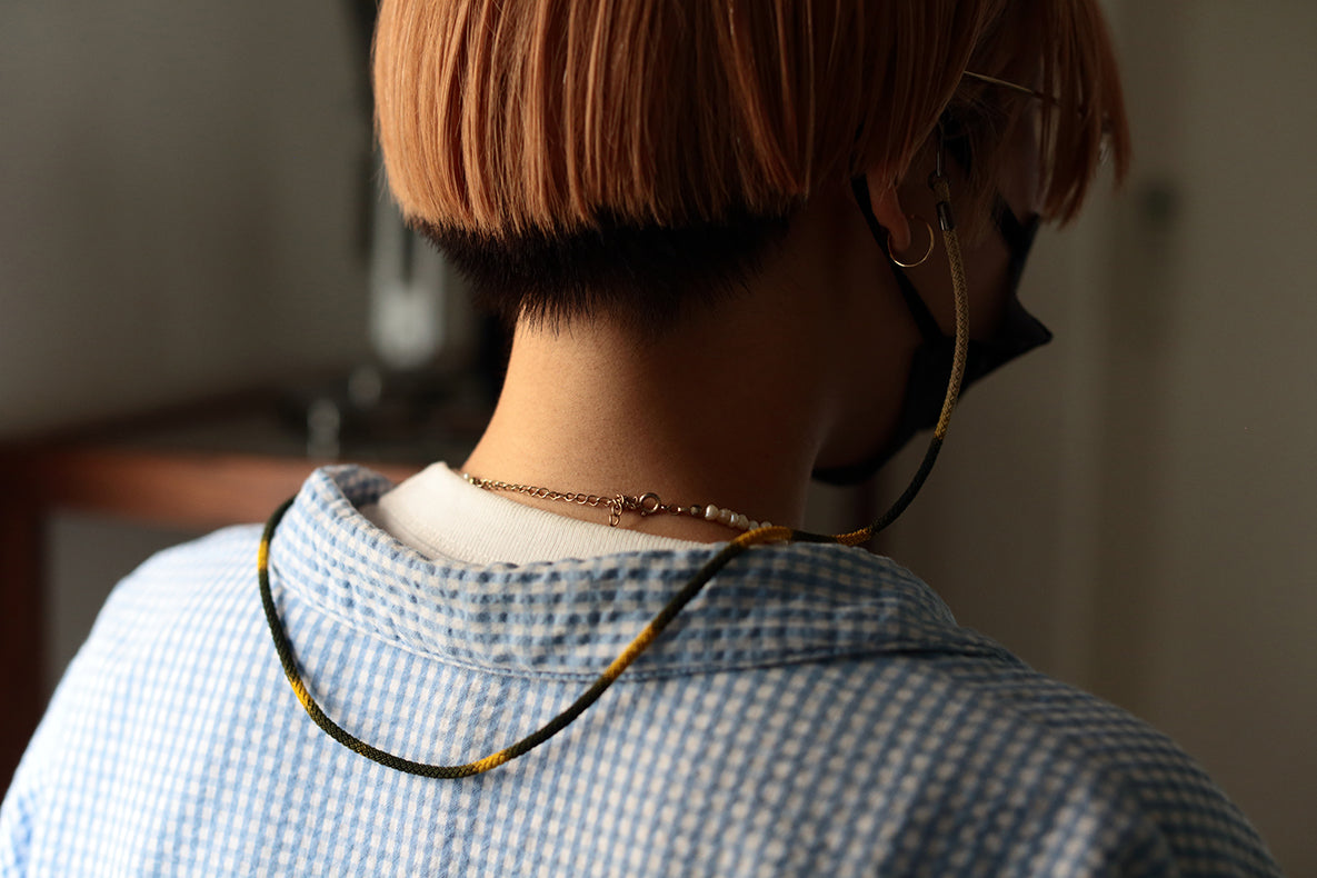A person with short, light brown hair wearing a black mask and glasses is seen from behind. They are wearing a blue checkered shirt and a beaded necklace featuring the DIFFUSER STRIPE DYEING GLASS CODE by DIFFUSER, which serves as a stylish glasses strap made from Japanese polyester cord and chain.