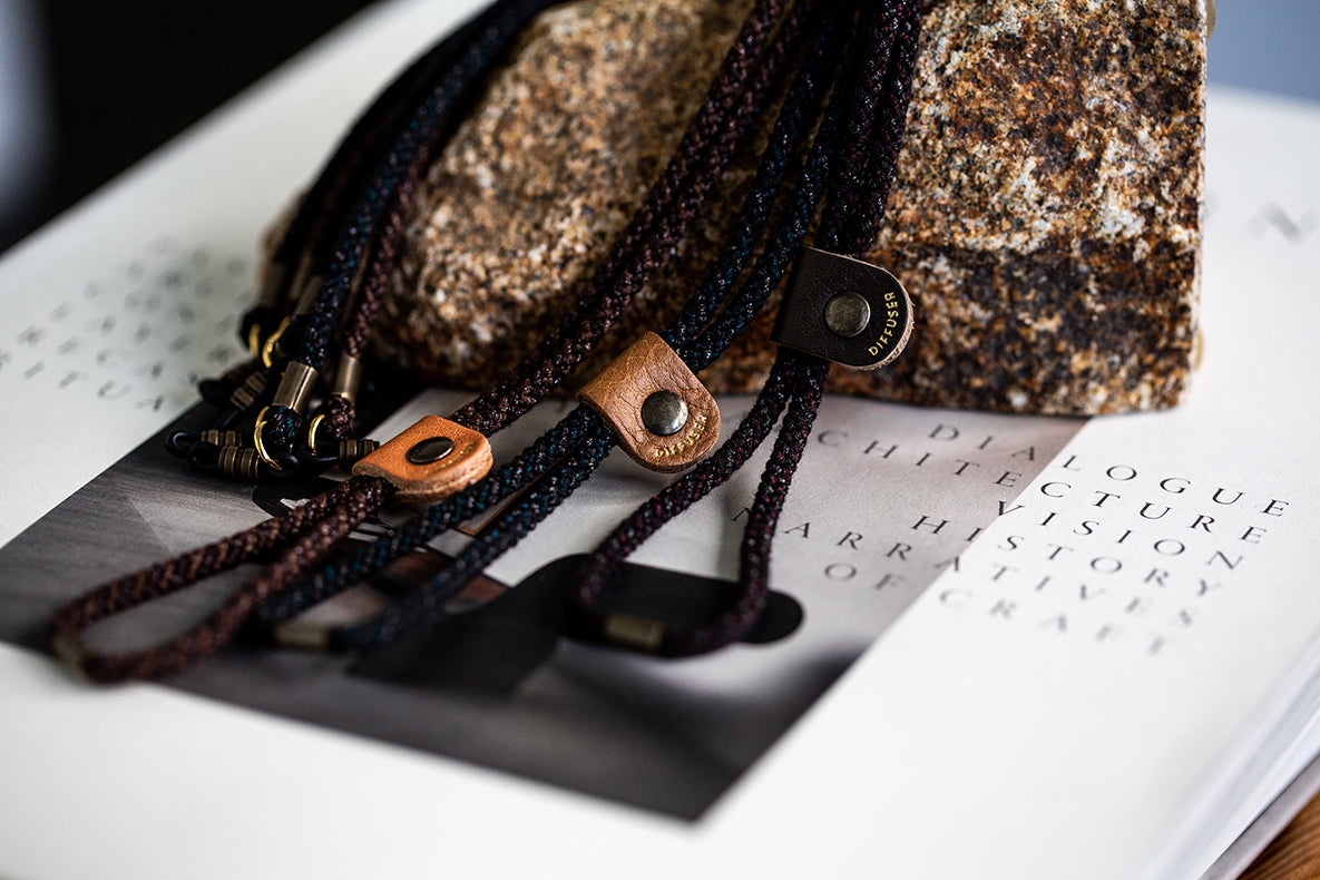 Close-up of braided black and brown cords with leather loops and metal snap closures, arranged on top of a printed book page and a rough stone.