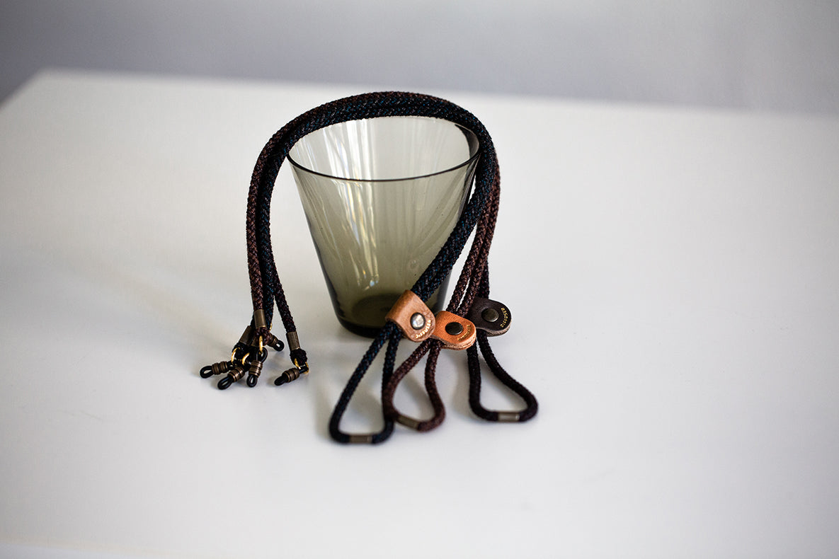 A translucent smoky amber glass tumbler with a dark brown braided strap and leather loop closure, photographed on a clean white surface.