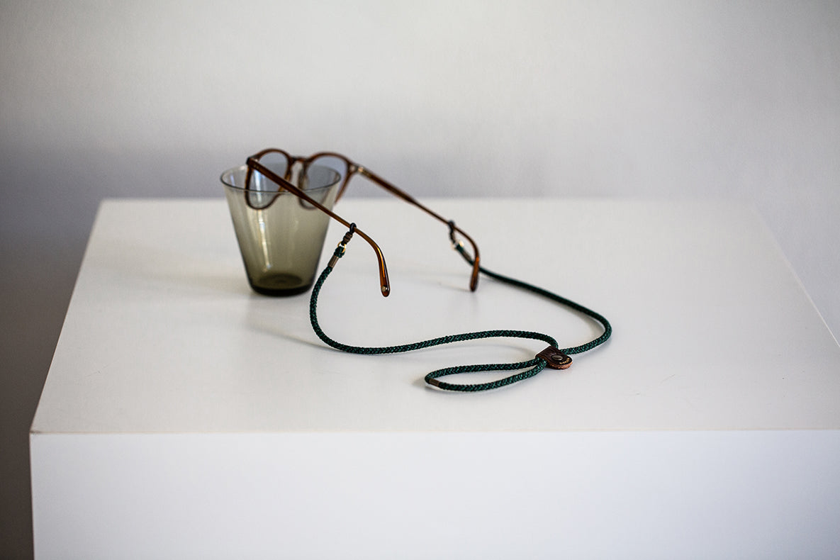 Retro-style eyeglasses with a dark green braided neck strap, resting on a white display block next to a small smoky glass cup.