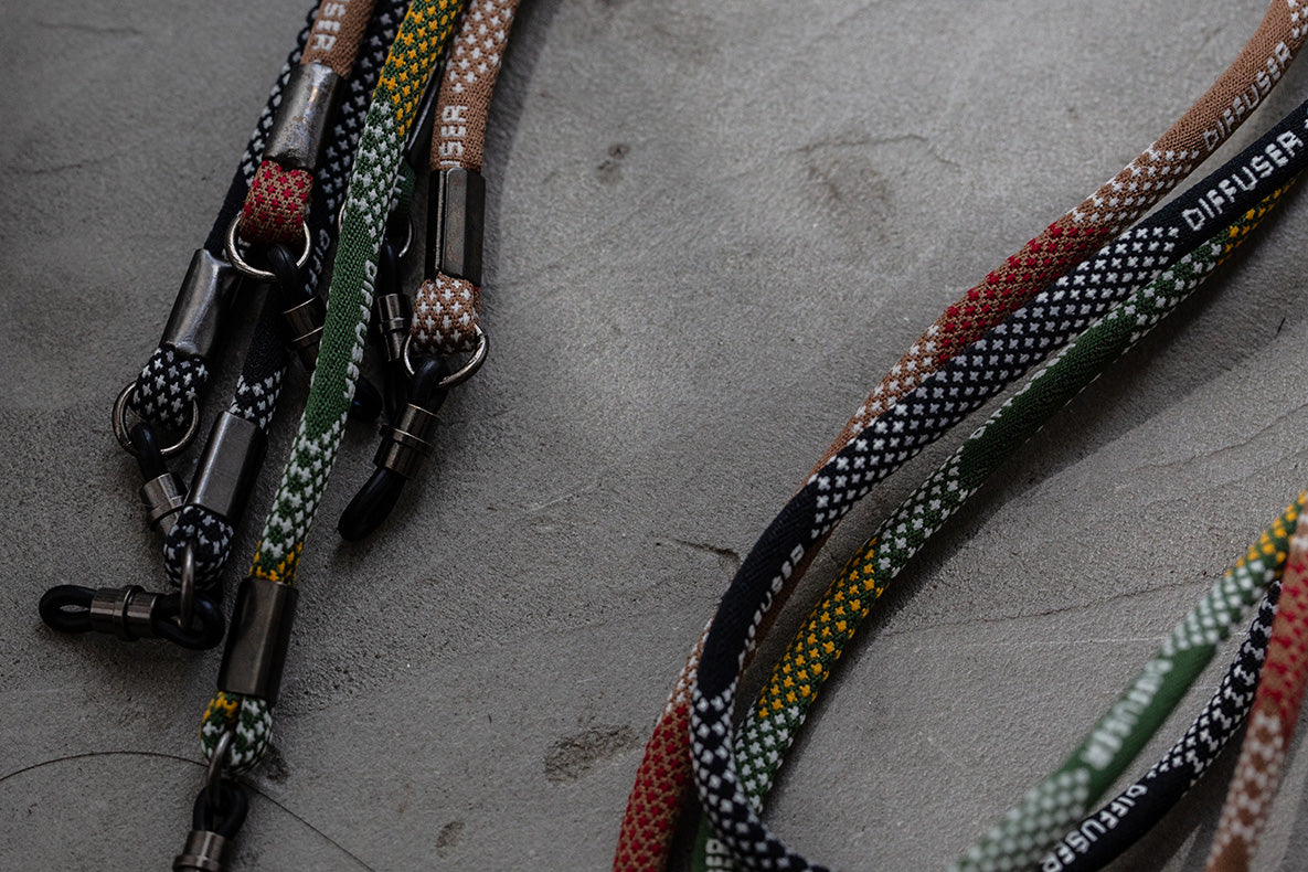 Several woven rope lanyards with metal clasps and rings laid out on a gray concrete surface. The lanyards feature multicolor patterns including black, white, green, red, yellow, and brown, with some showing the brand name 'DIFUSER'.