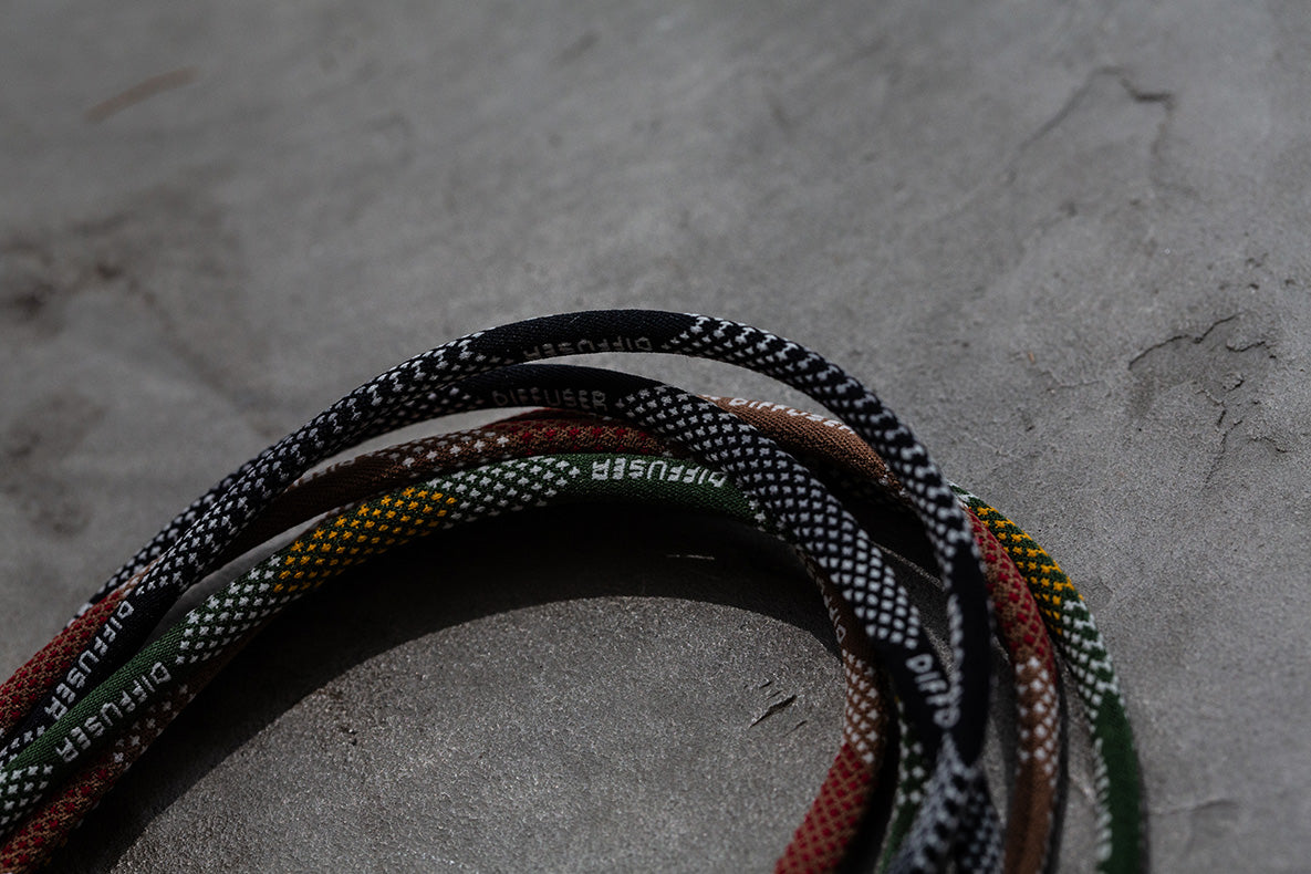 A close-up of several braided cords or straps coiled on a textured gray surface. The cords show mixed colors including black, white, green, red, and yellow, with some white text branding visible on the braid.