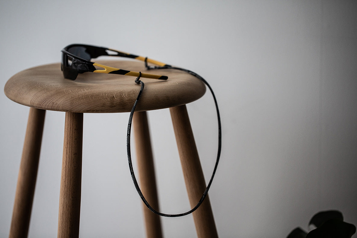 Black sunglasses with yellow accents and a black cord strap, resting on a round wooden stool against a plain light gray background.