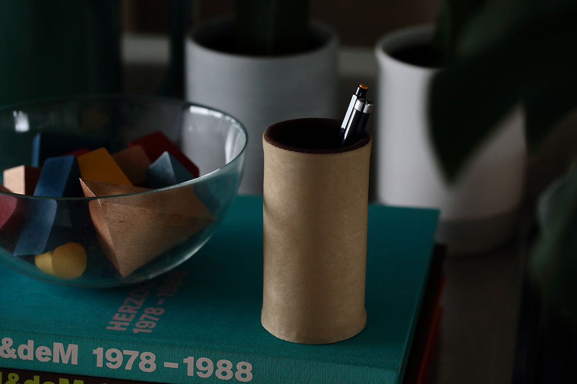 A black pen in a tan, suede-like nylon holder rests atop a teal book beside a glass bowl filled with colorful wooden blocks. Out-of-focus plants and pots create a soft background, while the DIFFUSER MILICLOTH EYEWEAR STAND by FRAMED EWE Optical—an elegant eyewear case crafted in Japan—adds a touch of sophistication to the scene.