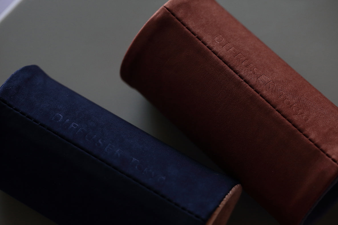Two cylindrical cases, one in navy blue and the other in brown, each with embossed “DIFFUSER TAG” text, are crafted from suede-like nylon as DIFFUSER MILICLOTH EYEWEAR STANDs by FRAMED EWE Optical. Made in Japan, these stylish eyewear cases rest elegantly atop a smooth, neutral-colored surface.