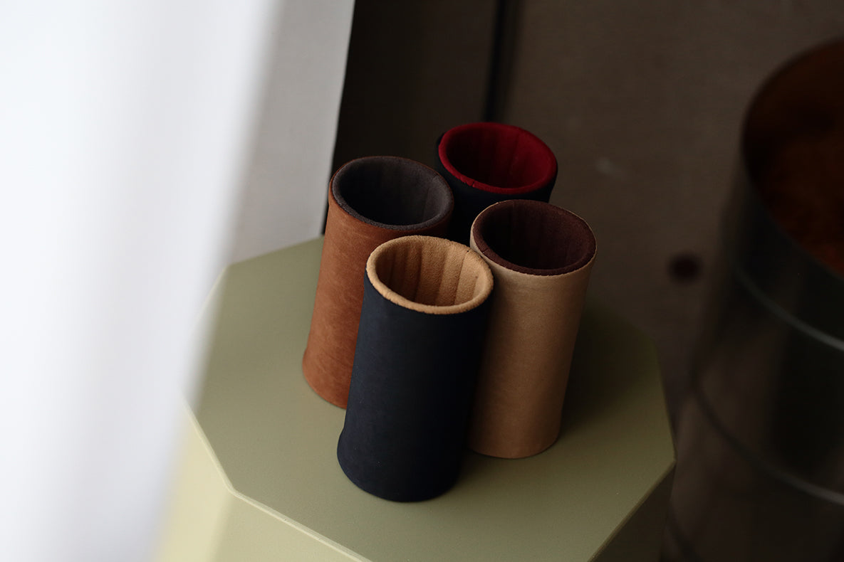 Four DIFFUSER MILICLOTH EYEWEAR STANDS by FRAMED EWE Optical—with suede-like nylon exteriors in black, brown, tan, and dark brown with a red interior—are arranged upright on a light green hexagonal surface near a window. Each stand is crafted in Japan.