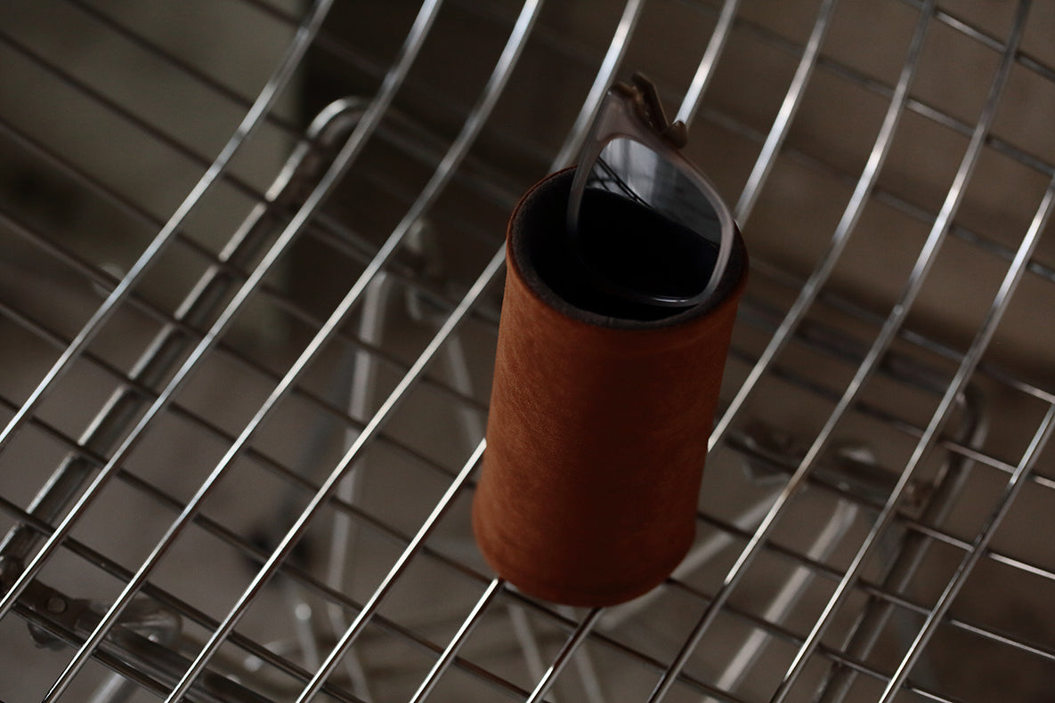 A DIFFUSER MILICLOTH EYEWEAR STAND by FRAMED EWE Optical, reminiscent of an elegant eyewear case with its brown leather sleeve, sits upright on a metallic wire grid surface.