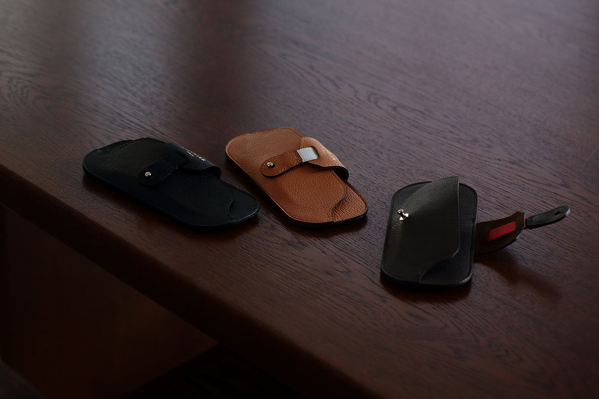 Three DIFFUSER SHRINK LEATHER SMART EYEWEAR CASES by DIFFUSER—crafted from Japanese cow leather and available in black, brown, and black with a red tag—are arranged in a row on a dark wooden surface. Each elegant case is made in Japan, showcasing the subtle refinement of traditional eyewear case design.