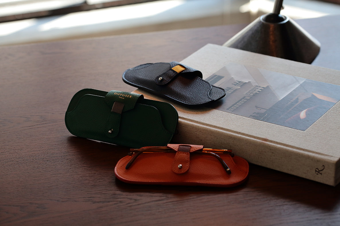 Three DIFFUSER SHRINK LEATHER SMART EYEWEAR CASES—in black, green, and brown—crafted from exquisite Japanese cow leather by DIFFUSER, are arranged on a book and wooden table. A pair of glasses is partially visible in the brown case. Soft natural light illuminates the scene, highlighting the refined craftsmanship made in Japan.