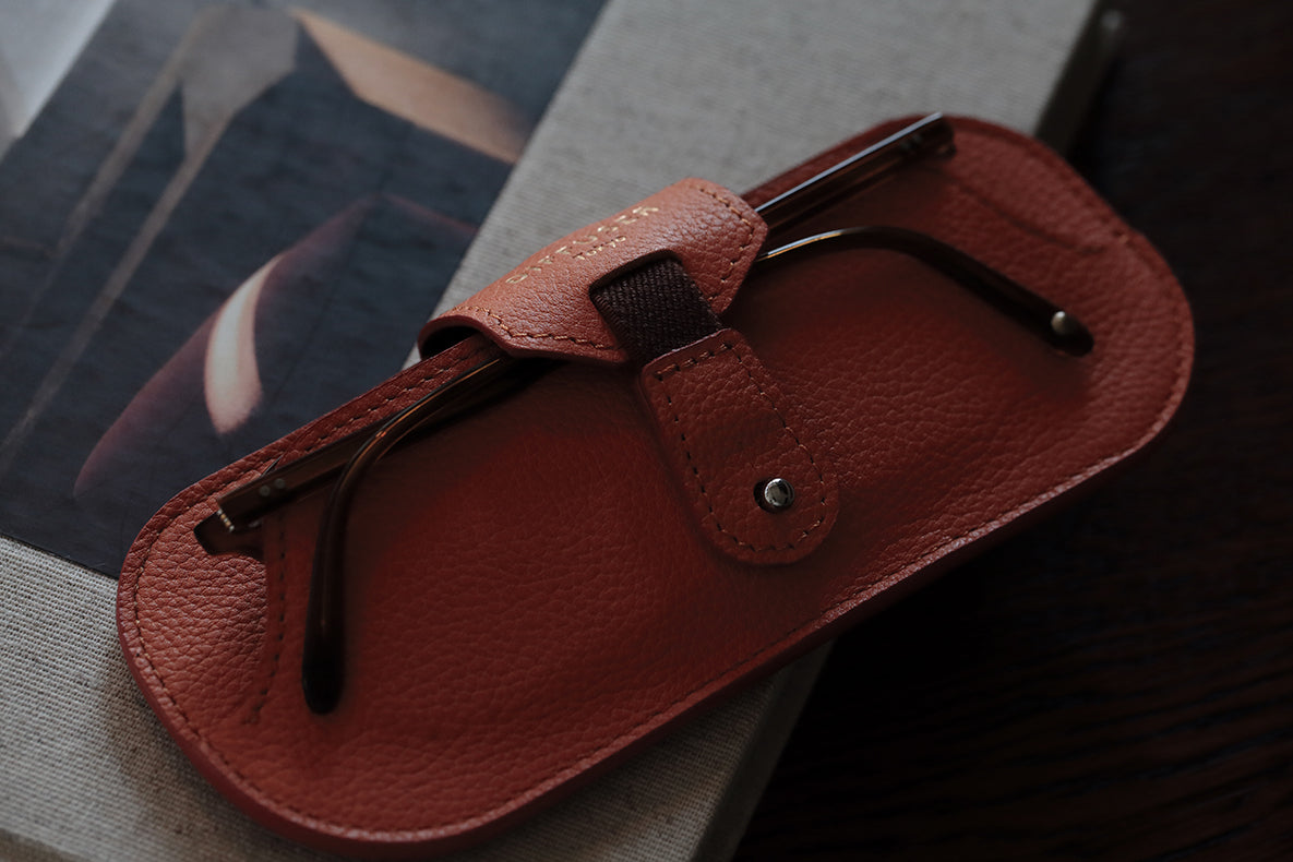 The DIFFUSER SHRINK LEATHER SMART EYEWEAR CASE by DIFFUSER, crafted from premium Japanese cow leather in brown, securely holds a pair of eyeglasses and is shown resting atop a closed book with an abstract-patterned cover peeking out.