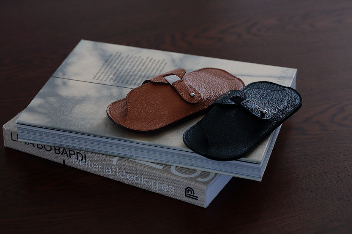 Two DIFFUSER SHRINK LEATHER SMART EYEWEAR CASES—one brown and one black, crafted from premium Japanese cow leather by DIFFUSER—rest on a closed book titled *Material Ideologies* atop a dark wooden surface.