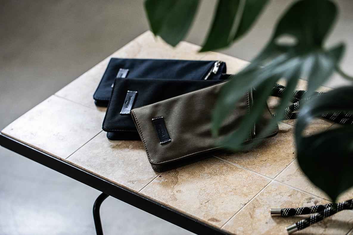 Three small zip pouches or wallet-style accessories in black, dark olive, and brown tones arranged on a tiled tabletop, with large green plant leaves partially framing the foreground.
