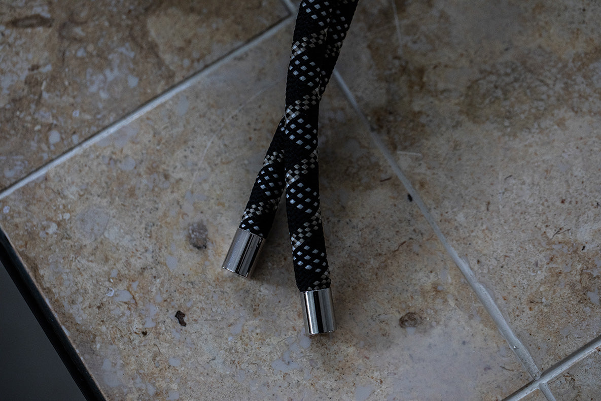 Black and white patterned braided laces with silver metal tips resting on a beige stone tile floor.