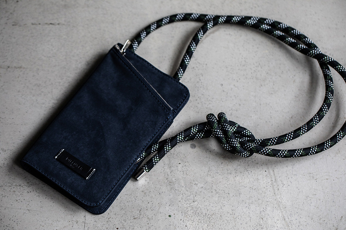 A dark navy blue suede pouch with a black and white speckled braided rope strap, featuring a silver-tone metal logo plate that reads 'SILKSTITCH'. The pouch is laid flat on a textured light gray concrete surface.