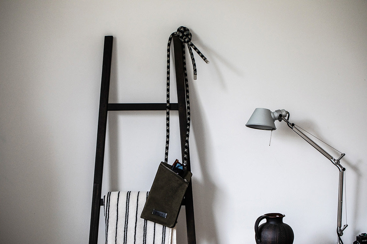 A black leather shoulder bag with a black-and-white striped strap hangs from a black ladder-style wall hook against a white wall, with a silver desk lamp and a dark ceramic vase nearby.