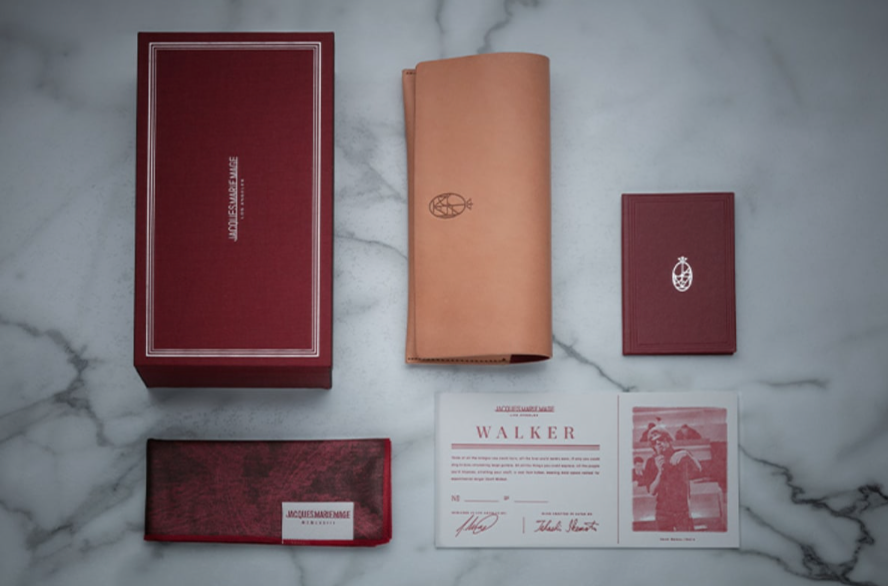Jacques Marie Mage Rimbaud box set displayed on a marble surface, featuring a red box, tan leather pouch, booklet, fabric case, certificate, and photo card—all adorned with the Jacques Marie Mage branding and logo—a collector’s must-have for those drawn to indie sleaze style and an appreciation for limited-edition spectacles.