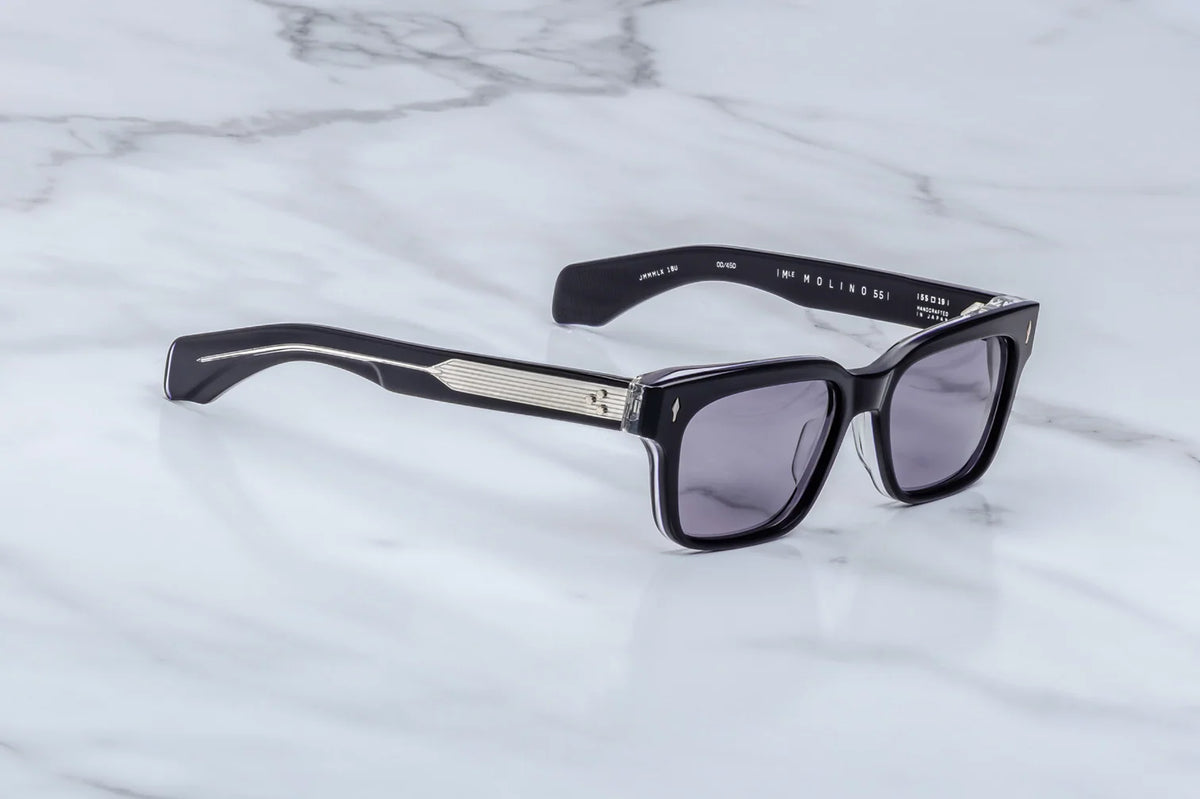 The Jacques Marie Mage Molino 55 sunglasses by Jacques Marie Mage, featuring a black rectangular frame with dark lenses, silver detailing on the temples, and subtle bevel accents, rest elegantly on a white marble surface.