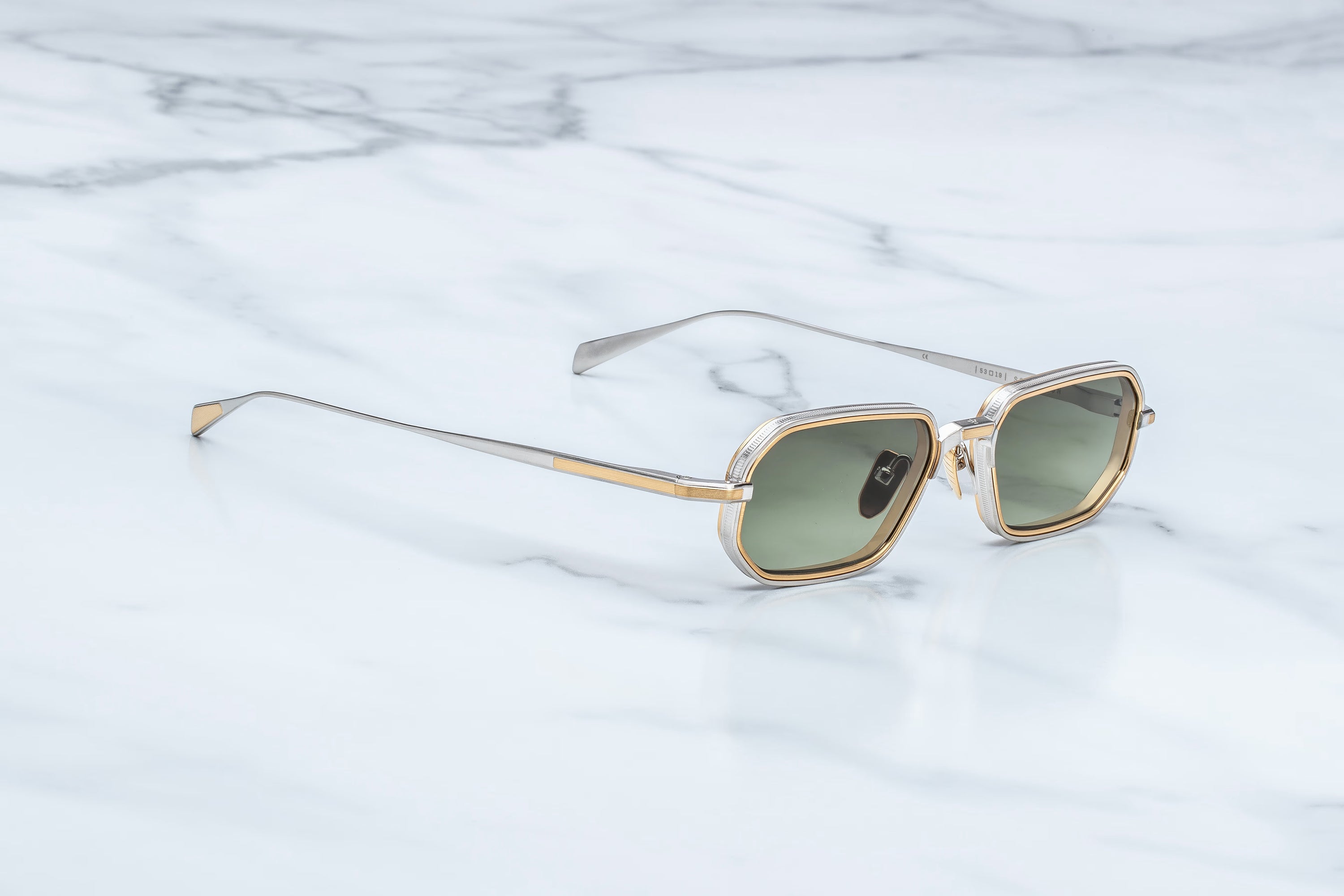 A pair of angular square sunglasses with a gold and silver-tone metal frame and green-tinted lenses, resting on a white marble surface.