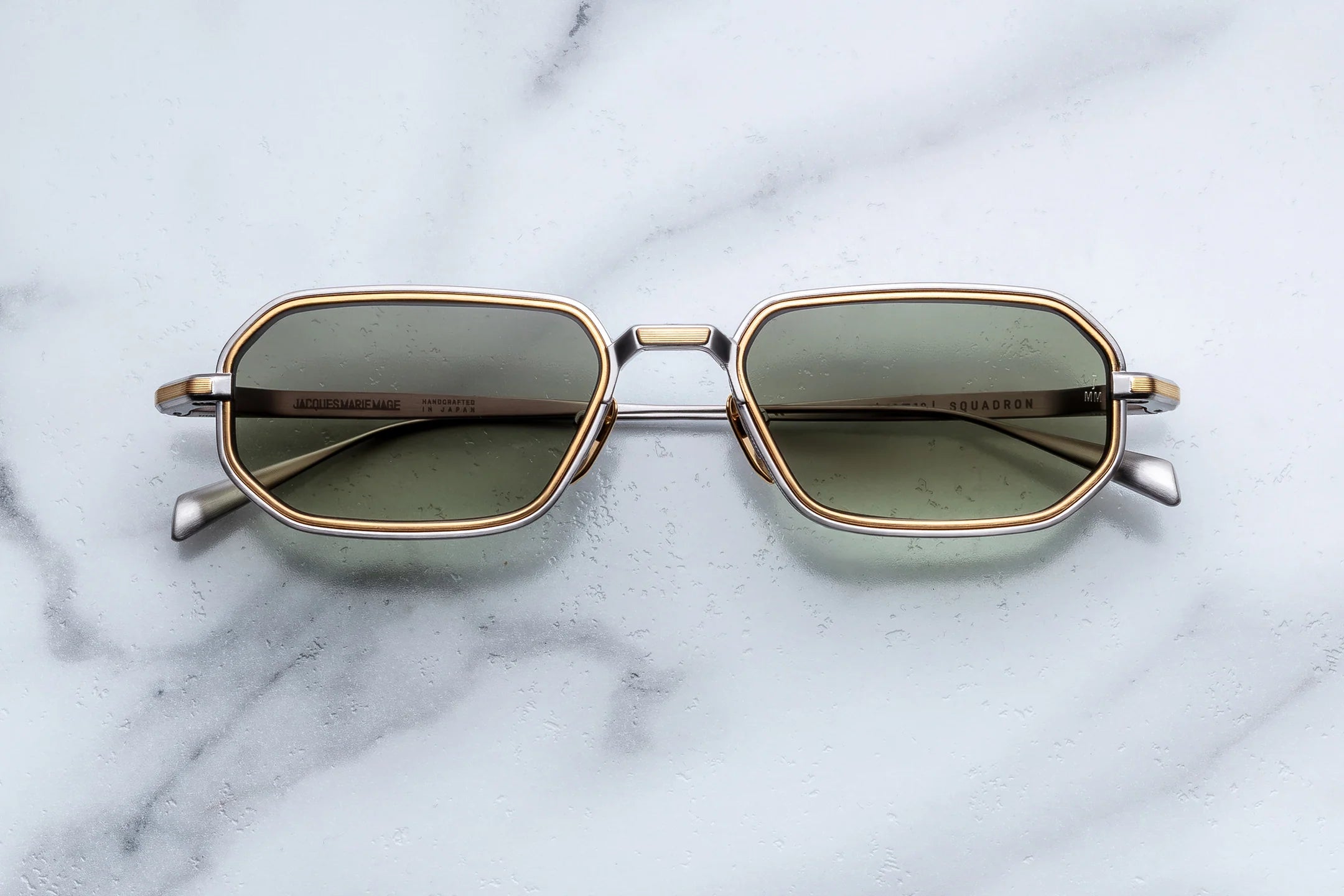 A pair of square-shaped sunglasses with a gold-tone metal frame and olive green lenses, resting on a white marble surface.