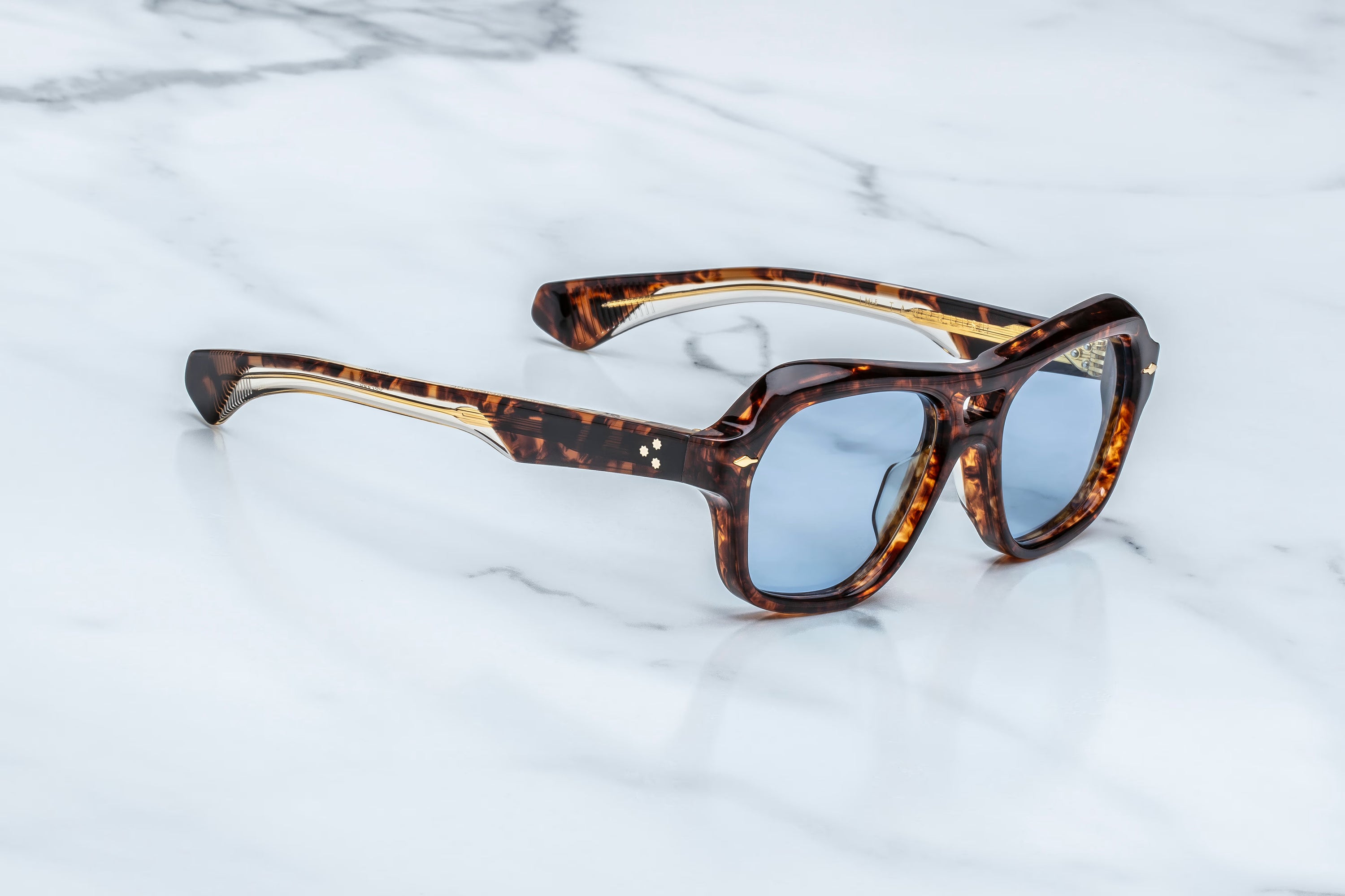 Showcasing a daring silhouette, the Jacques Marie Mage Tamerlis sunglasses by Jacques Marie Mage feature sculptural temples and blue-tinted lenses in a tortoiseshell frame, artfully displayed on a white marble surface.