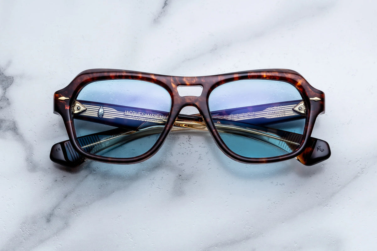 A pair of Jacques Marie Mage Tamerlis limited-edition spectacles in brown tortoiseshell with blue lenses, placed on a white marble surface. The daring silhouette and crossed arms are accentuated by subtle reflections.