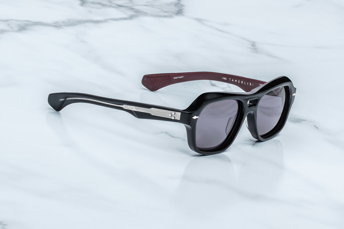 Jacques Marie Mage Tamerlis sunglasses with thick black frames and dark lenses rest on a white marble surface. Showcasing a daring silhouette, these limited-edition Jacques Marie Mage spectacles reveal sculptural temples in dark red on the inside, with text visible on the inner left temple.